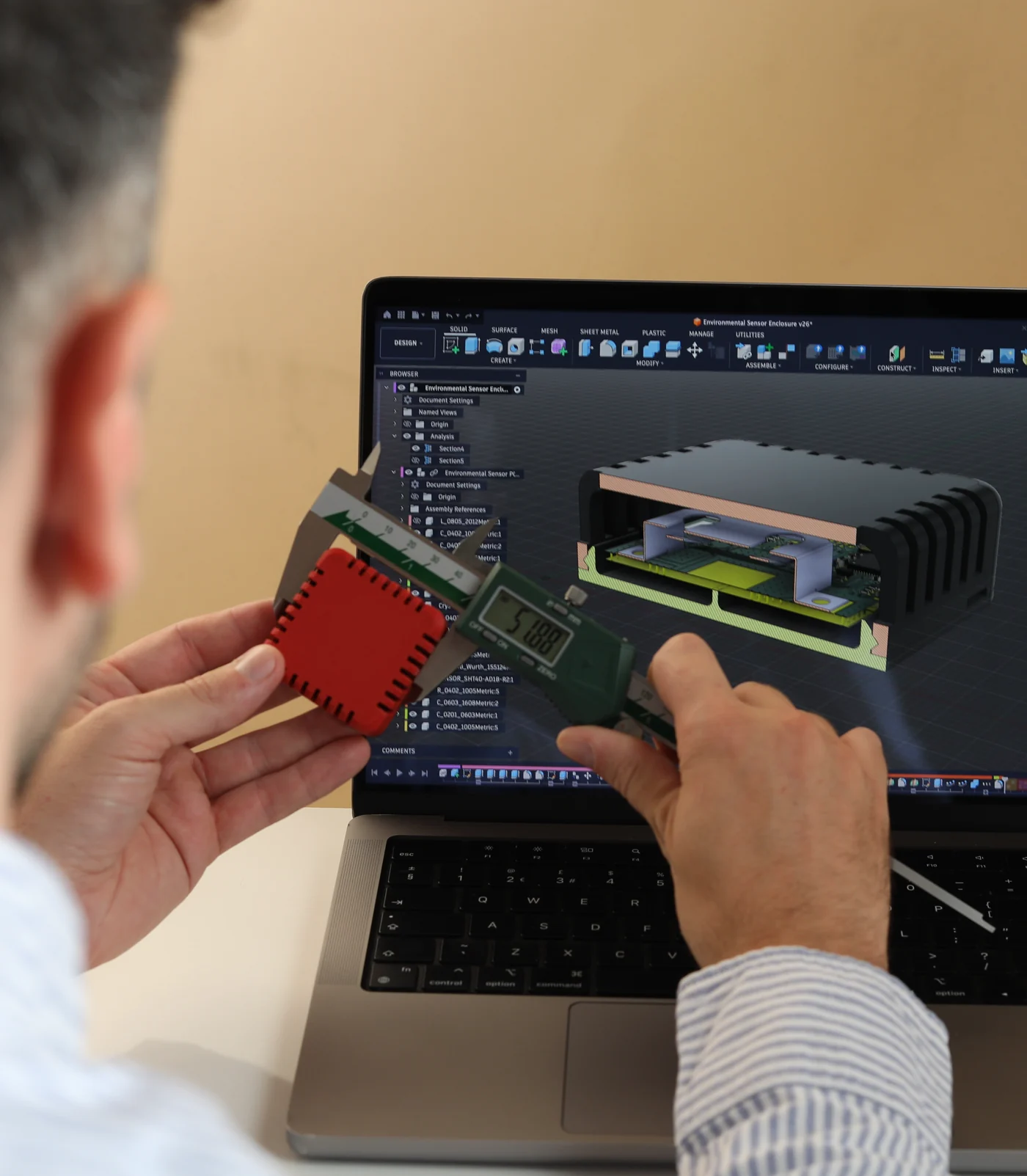 A Bitmask engineer measuring a 3D-printed enclosure with digital calipers, its CAD model open on the laptop behind
