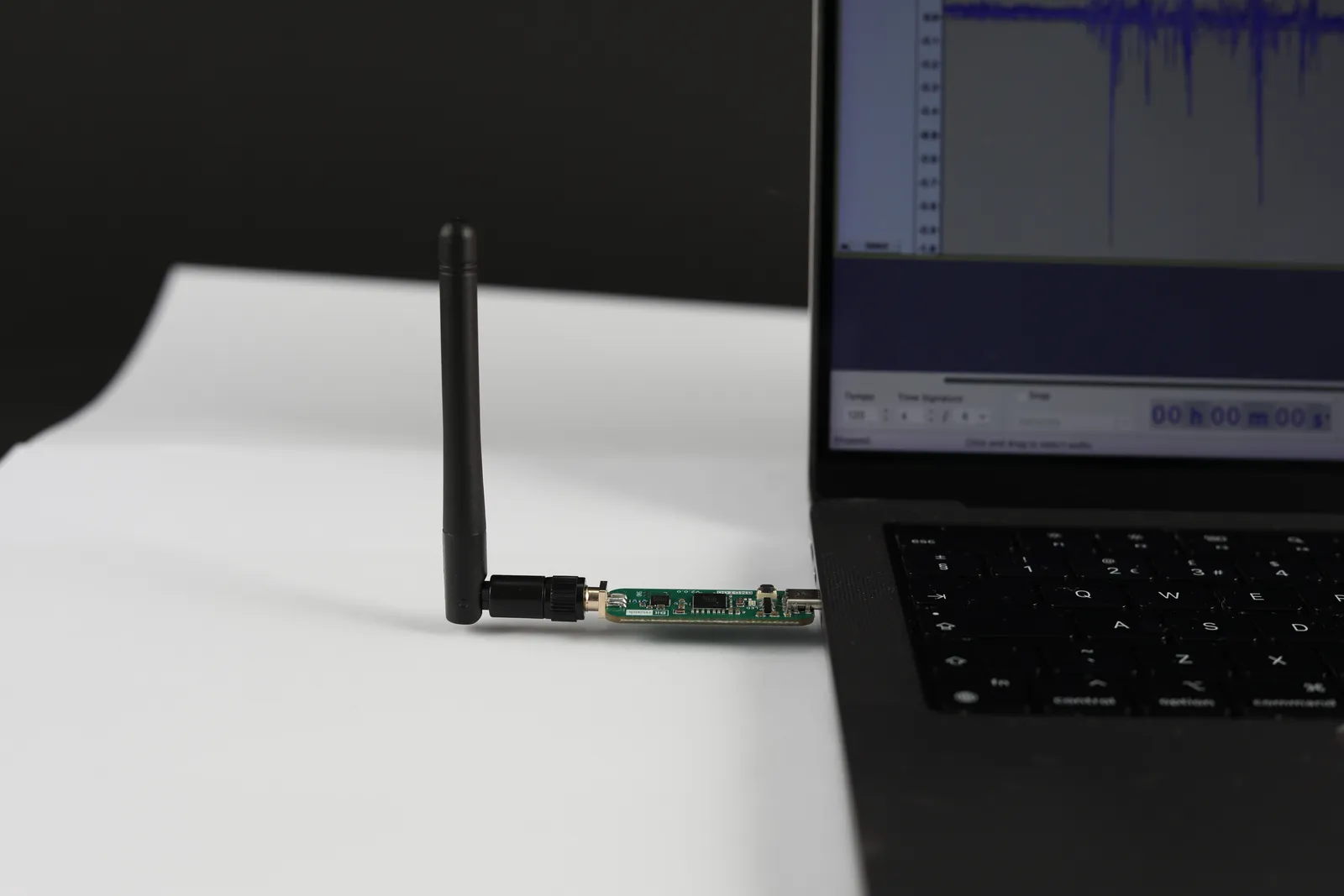 BMD100 dongle plugged into a laptop's USB-C port with its antenna fitted, an audio waveform on the screen behind it